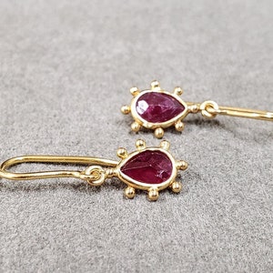 May include: A pair of gold earrings featuring teardrop-shaped ruby stones. The earrings have a delicate design with small gold beads surrounding the stones.