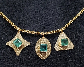 May include: A gold necklace with three pendants, each featuring a different geometric shape: a diamond, a circle, and a triangle. Each pendant has a green emerald gemstone in the centre.