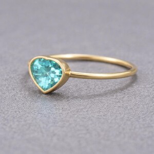May include: A gold ring with a heart-shaped, light blue gemstone. The ring's band is thin and gold, and the gemstone is set in a gold bezel. The ring is displayed on a gray surface. Jewelry.