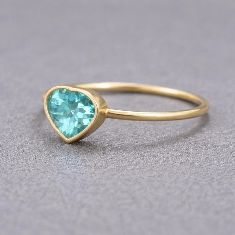 May include: A gold ring with a heart-shaped, light blue gemstone. The ring's band is thin and gold, and the gemstone is set in a gold bezel. The ring is displayed on a gray surface. Jewelry.