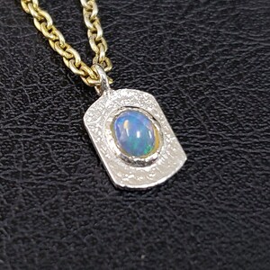 May include: A silver pendant necklace with a textured finish and a blue opal gemstone set in a silver bezel. The necklace is on a gold chain.