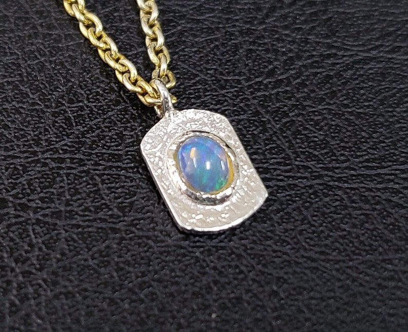 May include: A silver pendant necklace with a textured finish and a blue opal gemstone set in a silver bezel. The necklace is on a gold chain.