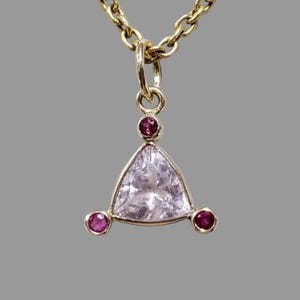 May include: A gold necklace with a triangular pendant. The pendant has a large, clear, triangular-cut stone, with three smaller, round, red gemstones. The chain is gold, creating a cohesive design. The necklace is suitable for everyday wear or special occasions.