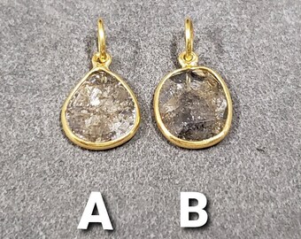 Puede incluir: Dos pendientes colgantes dorados. Uno tiene forma de lágrima, el otro es ovalado. Ambos tienen una piedra rugosa, gris y transparente engastada en un bisel dorado. Los pendientes se muestran sobre una superficie gris. La letra "A" está debajo del pendiente en forma de lágrima, y la letra "B" debajo del pendiente ovalado.