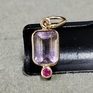 May include: A gold pendant with a large, rectangular, purple amethyst gemstone and a smaller, round, pink ruby gemstone.