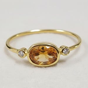 Può includere: Un anello d'oro con una gemma ovale color ambra al centro, affiancata da due gemme rotonde e chiare più piccole. L'anello ha una sottile fascia d'oro lucido. Le gemme sono incastonate in una montatura a castone.