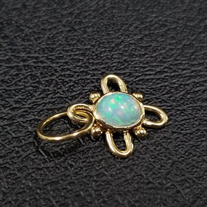 May include: A gold charm with a white opal gemstone in the center. The charm is shaped like a flower with four petals and has small gold beads around the opal.
