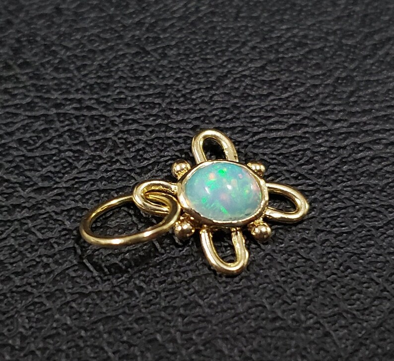 May include: A gold charm with a white opal gemstone in the center. The charm is shaped like a flower with four petals and has small gold beads around the opal.