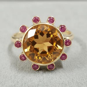 May include: A gold ring featuring a large, round, faceted, amber-colored gemstone at the center, surrounded by smaller, circular, pink gemstones. The ring's band is a simple, polished gold.