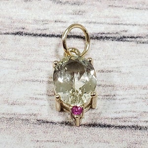 May include: Gold pendant featuring a large, oval-cut, pale green gemstone with a smaller, heart-shaped, pink gemstone below. The pendant has a gold bail for a chain.
