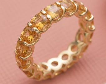 May include: A gold ring featuring a continuous row of rectangular, amber-colored gemstones. The ring's band is also gold, with a delicate, openwork design. The gemstones are set closely together, creating a seamless, sparkling effect. The ring is displayed against a soft pink background.