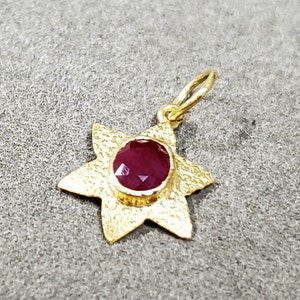 May include: A gold-colored pendant in the shape of a star with six points. The center of the star holds a round, deep red gemstone. The pendant has a textured surface and a small loop for a chain.