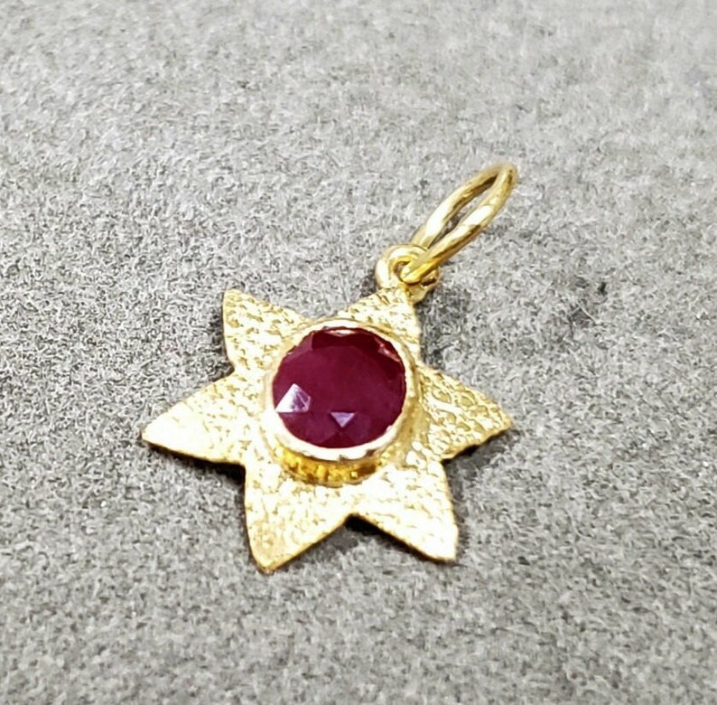 May include: A gold-colored pendant in the shape of a star with six points. The center of the star holds a round, deep red gemstone. The pendant has a textured surface and a small loop for a chain.
