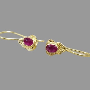 Puede incluir: Un par de pendientes dorados con diseño de gancho. Cada pendiente presenta una vibrante gema de color rubí ovalada, engastada en un marco dorado decorativo y texturizado. Los pendientes se muestran sobre un fondo gris.