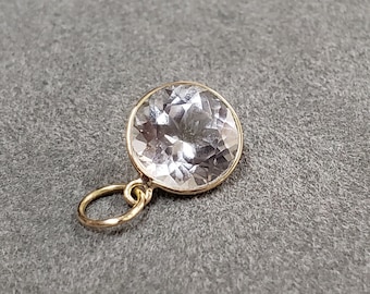 May include: A round, clear gemstone pendant with a gold-colored bezel and loop. The gemstone has multiple facets, reflecting light. The pendant is set against a gray background.