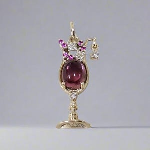 May include: A gold cocktail pendant featuring a large, oval, deep red gemstone. The pendant is embellished with smaller pink and clear gemstones, and a small dangling charm. The design is reminiscent of a cocktail glass.