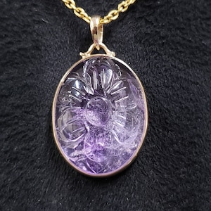 Op de afbeelding: Een hanger van paarse amethyst met een gegraveerd bloemmotief, bevestigd aan een gouden ketting.