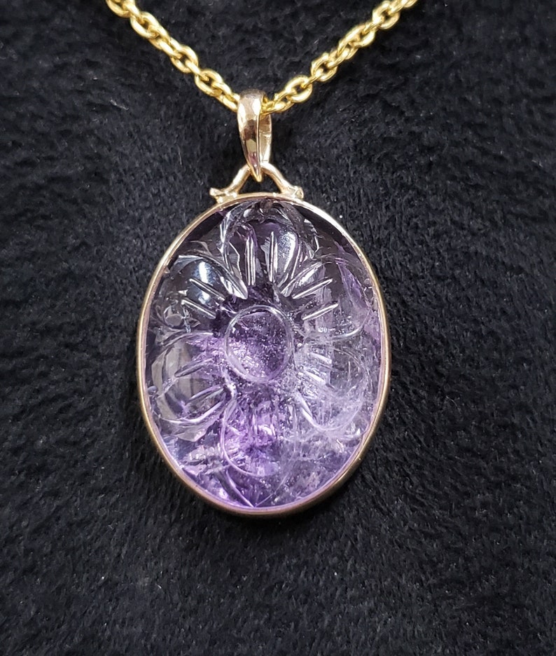 Op de afbeelding: Een hanger van paarse amethyst met een gegraveerd bloemmotief, bevestigd aan een gouden ketting.