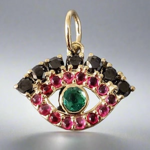 Puede incluir: Un amuleto de oro en forma de ojo con piedras preciosas negras y rojas que rodean una piedra preciosa verde en el centro.
