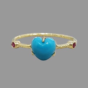 May include: A gold ring featuring a central heart-shaped turquoise stone held by four prongs. The band has a textured, hammered finish and is accented on each side by a small, round, deep red gemstone set into the metal shank of the ring.