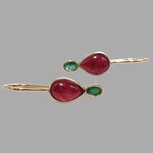 May include: A pair of gold earrings featuring a teardrop-shaped, deep red gemstone paired with a smaller, oval-shaped green gemstone. The earrings have a simple, elegant design.