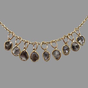 May include: A gold necklace with a fine chain, adorned with ten irregularly shaped, clear-grey gemstone pendants. Each pendant is suspended from the chain by a small gold ring. The gemstones have a speckled appearance.