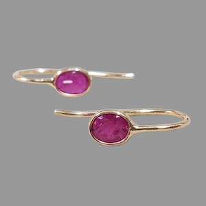 Puede incluir: Dos pendientes dorados, cada uno con una vibrante gema de color magenta de forma ovalada. Los pendientes tienen un diseño simple y elegante, con un gancho curvado para la oreja. El fondo es gris neutro.