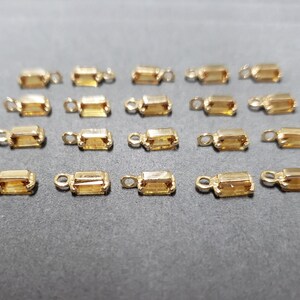 May include: A collection of small, rectangular gold-colored jewelry findings with a loop at one end and a rectangular, amber-colored stone in the center. The findings are arranged on a dark gray surface.