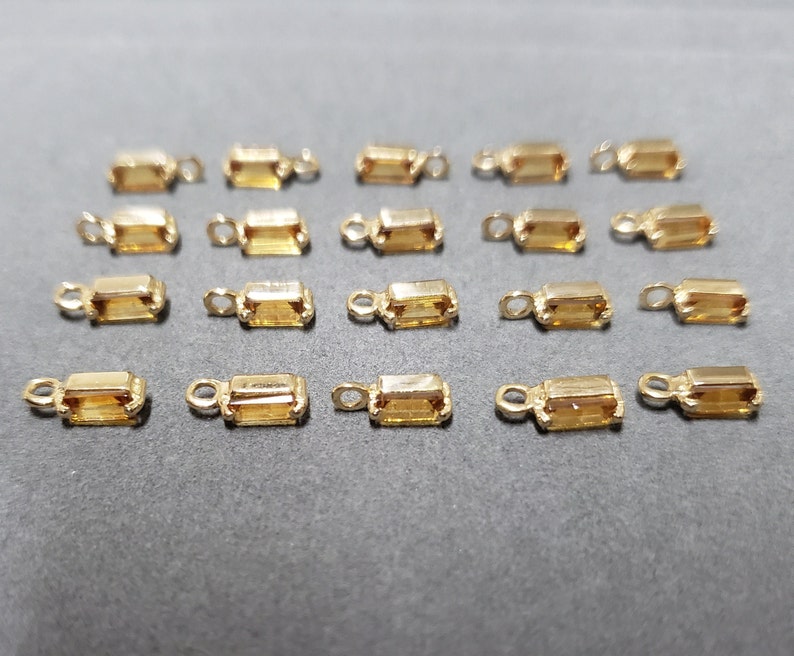 May include: A collection of small, rectangular gold-colored jewelry findings with a loop at one end and a rectangular, amber-colored stone in the center. The findings are arranged on a dark gray surface.
