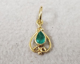 May include: A gold-colored pendant with a teardrop-shaped emerald gemstone. The pendant has a decorative, openwork design with a loop at the top for hanging. The emerald is a vibrant green color.