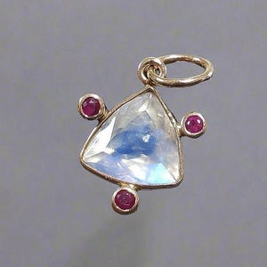 Könnte beinhalten: Goldfarbener Anhänger mit einem dreieckigen, schillernden Mondstein in der Mitte und drei kleinen, runden, roten Edelsteinen. Der Anhänger hat oben eine Schlaufe für eine Kette. Das Design ist schlicht und elegant.