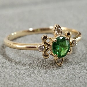 Puede incluir: Un anillo de oro con una piedra preciosa verde rodeada de diamantes más pequeños. El anillo tiene un diseño delicado e intrincado.