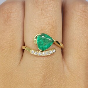 May include: A gold ring featuring a pear-shaped, vibrant green emerald gemstone. The ring has a curved band with small, clear gemstones set along its length. The emerald is the focal point, set in a unique, elegant design.