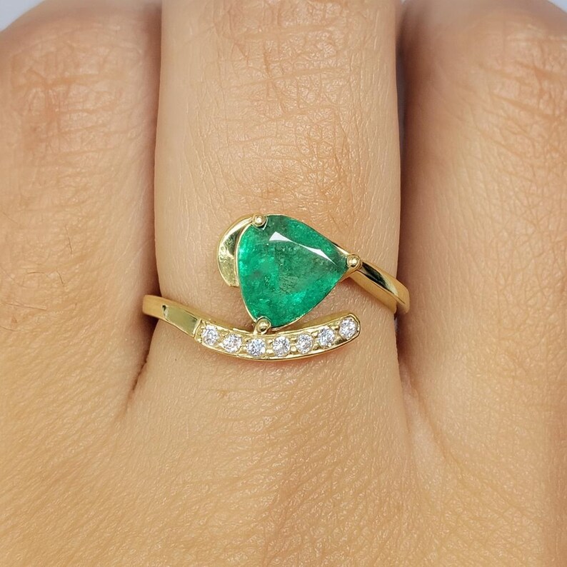 May include: A gold ring featuring a pear-shaped, vibrant green emerald gemstone. The ring has a curved band with small, clear gemstones set along its length. The emerald is the focal point, set in a unique, elegant design.