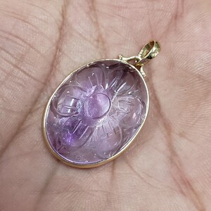 Op de afbeelding: Een hanger van paarse amethyst, gesneden met een bloemmotief, ingelijst in een gouden metalen frame.