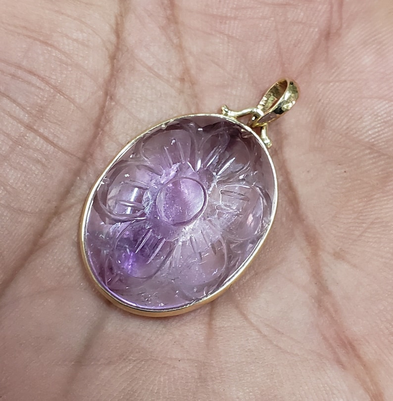 Op de afbeelding: Een hanger van paarse amethyst, gesneden met een bloemmotief, ingelijst in een gouden metalen frame.