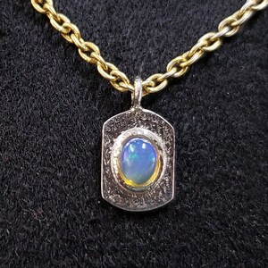 May include: A silver pendant necklace with a gold chain. The pendant features a textured silver frame with a blue opal gemstone in the center.