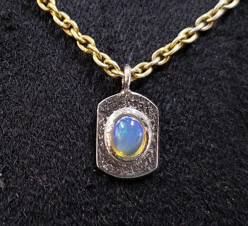 May include: A silver pendant necklace with a gold chain. The pendant features a textured silver frame with a blue opal gemstone in the center.