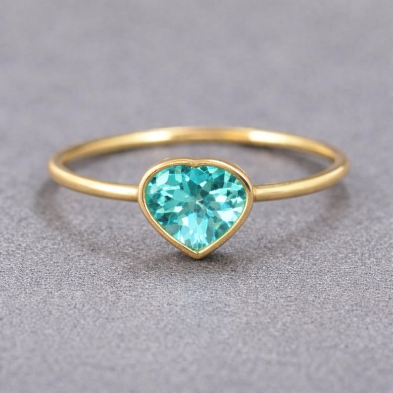 May include: A gold ring featuring a heart-shaped, light blue gemstone. The gemstone is set in a gold bezel, and the band is a thin, polished gold. The ring is displayed on a gray background.