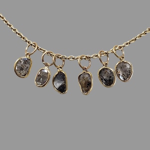 May include: A gold chain necklace featuring six dangling, irregular, raw-cut grey and black stone charms, each encased in a thin gold bezel setting. The stones vary in shape and opacity, suspended from small gold jump rings along the chain.