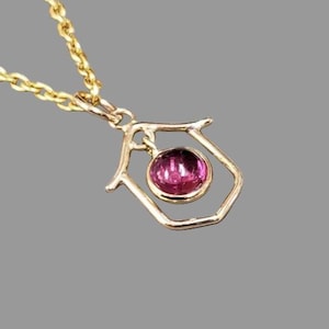 May include: A gold necklace with a pendant. The pendant has a magenta gemstone set within an open, geometric frame. The chain is made of small, linked segments. The gemstone is a vibrant, circular shape.