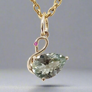 May include: A gold pendant necklace featuring a green gemstone in the shape of a teardrop, set in a swan design with a small pink gemstone accent.