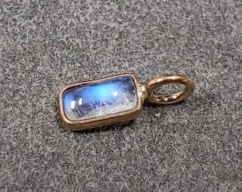 Könnte beinhalten: Ein goldener Anhänger mit einem rechteckigen, blauen Mondstein-Anhänger. Der Anhänger hat eine kleine Öse zum Anbringen an einer Halskette oder einem Armband.