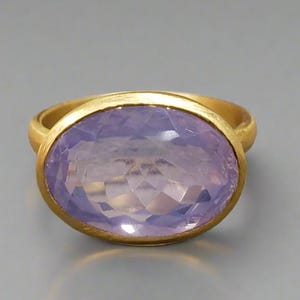 Puede incluir: Un anillo de oro con una gran piedra preciosa ovalada de color lila claro. La piedra preciosa está engastada en un bisel de oro, y la banda del anillo también es de oro. El fondo es de color gris claro.