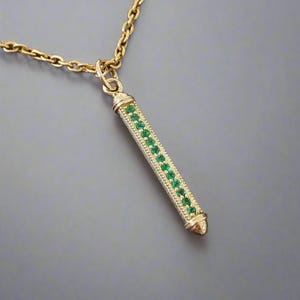 Puede incluir: Un collar dorado con un colgante de diseño rectangular. El colgante está adornado con una fila de piedras preciosas verdes. La cadena del collar también es dorada, creando un aspecto cohesivo y elegante. La joya se presenta sobre un fondo gris.