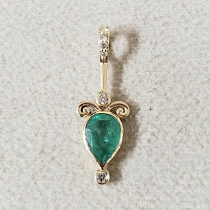 May include: A gold pendant with a pear-shaped emerald gemstone surrounded by small diamonds. The pendant has a delicate design with swirls and a bail at the top.