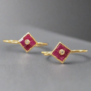 Puede incluir: Dos anillos de oro, cada uno con un engaste cuadrado de rubíes y un diamante central. Los rubíes son de color rojo intenso y las bandas son doradas. Los anillos se muestran sobre un fondo gris.