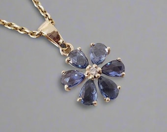 May include: A gold-toned necklace featuring a flower pendant. The pendant is composed of six pear-shaped, deep blue gemstones surrounding a central cluster of smaller, clear stones. The chain is a delicate, twisted design.