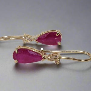 May include: A pair of gold drop earrings with pear-shaped, deep red gemstones. The earrings have a delicate design with small accent stones near the top of the drop. The gold has a polished finish, and the earrings are displayed on a neutral background.