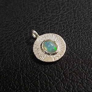 May include: A silver pendant with a round, textured frame and a light blue opal gemstone in the center.